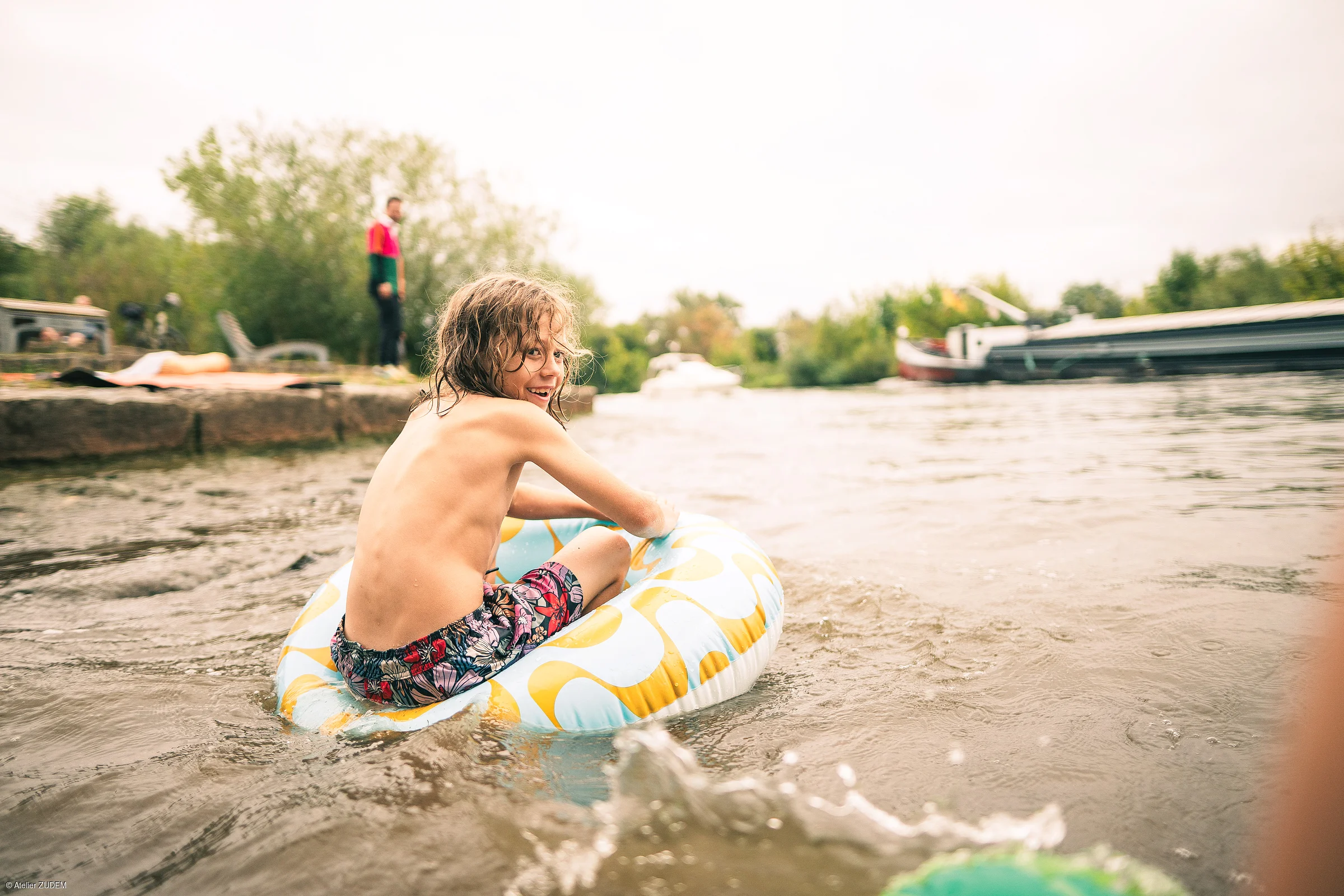 Kind sitzt in Schwimmring im Wasser, im Hintergrund Ufer mit stehender Person und Booten.