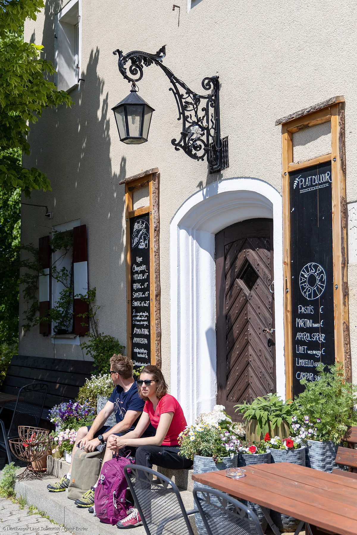 Zwei Personen sitzen vor einem Haus mit Holztür, Blumenkübeln und zwei Schiefertafeln mit Speisekarten.