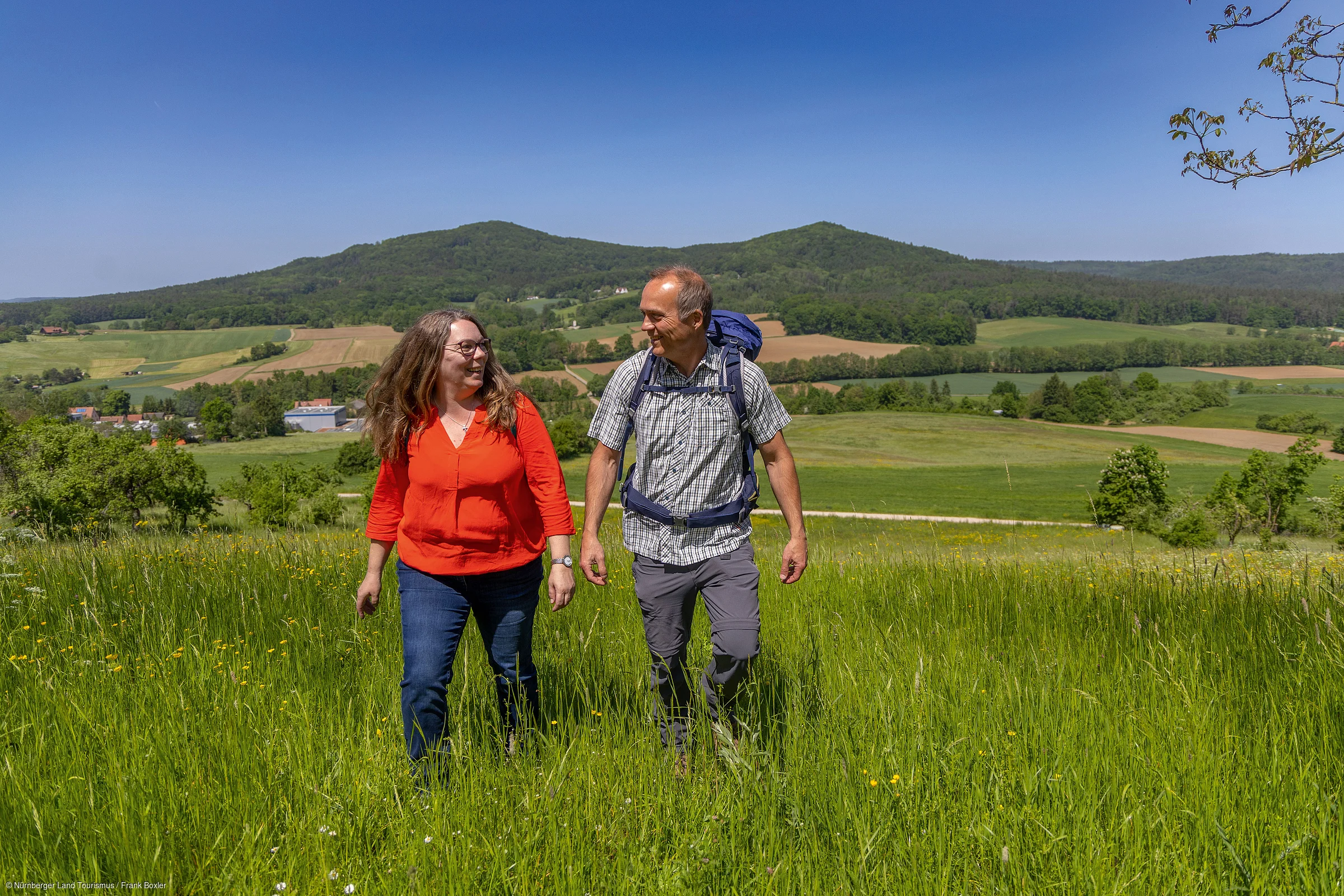 Zwei Personen wandern auf einer grünen Wiese mit Hügeln und Feldern im Hintergrund bei klarem Himmel.