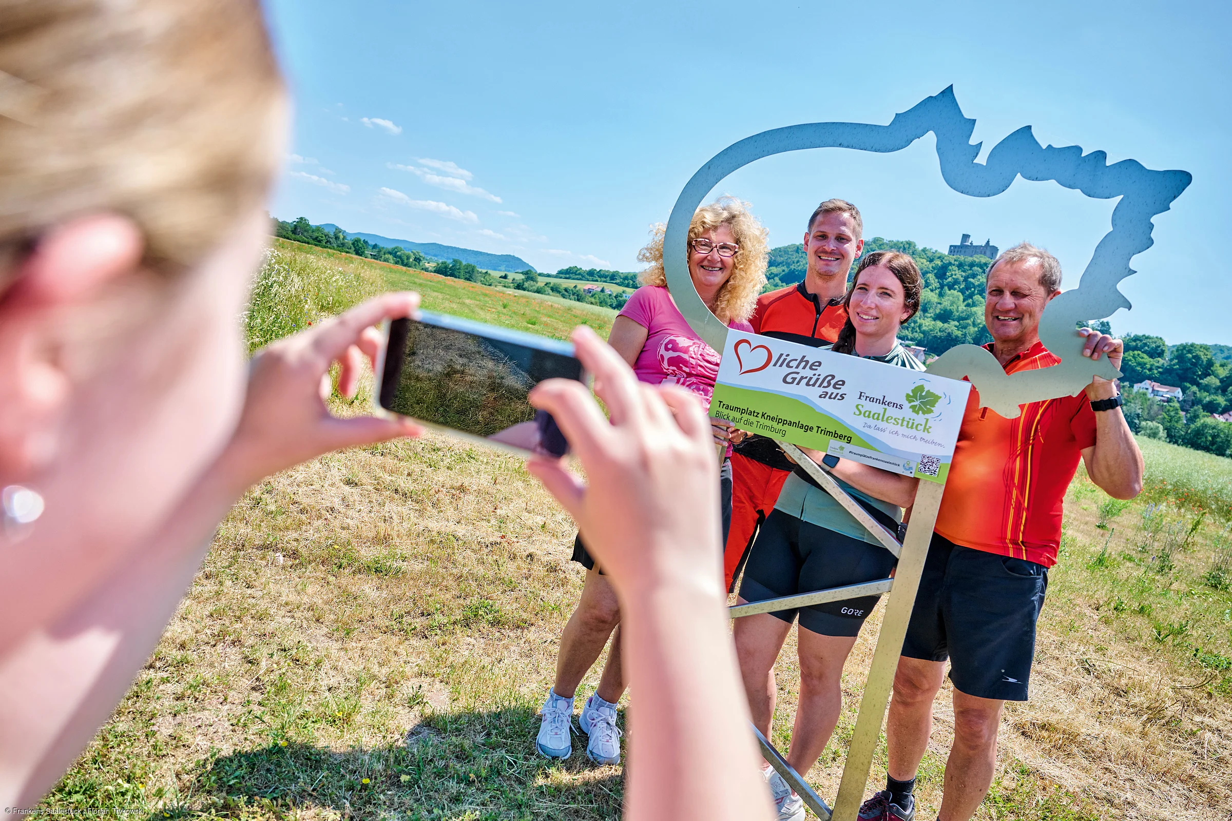 Vier Personen posieren mit einem Rahmen und Schild „Herzliche Grüße aus Frankens Saalestück“ auf einer Wiese, eine fünfte Person fotografiert sie.