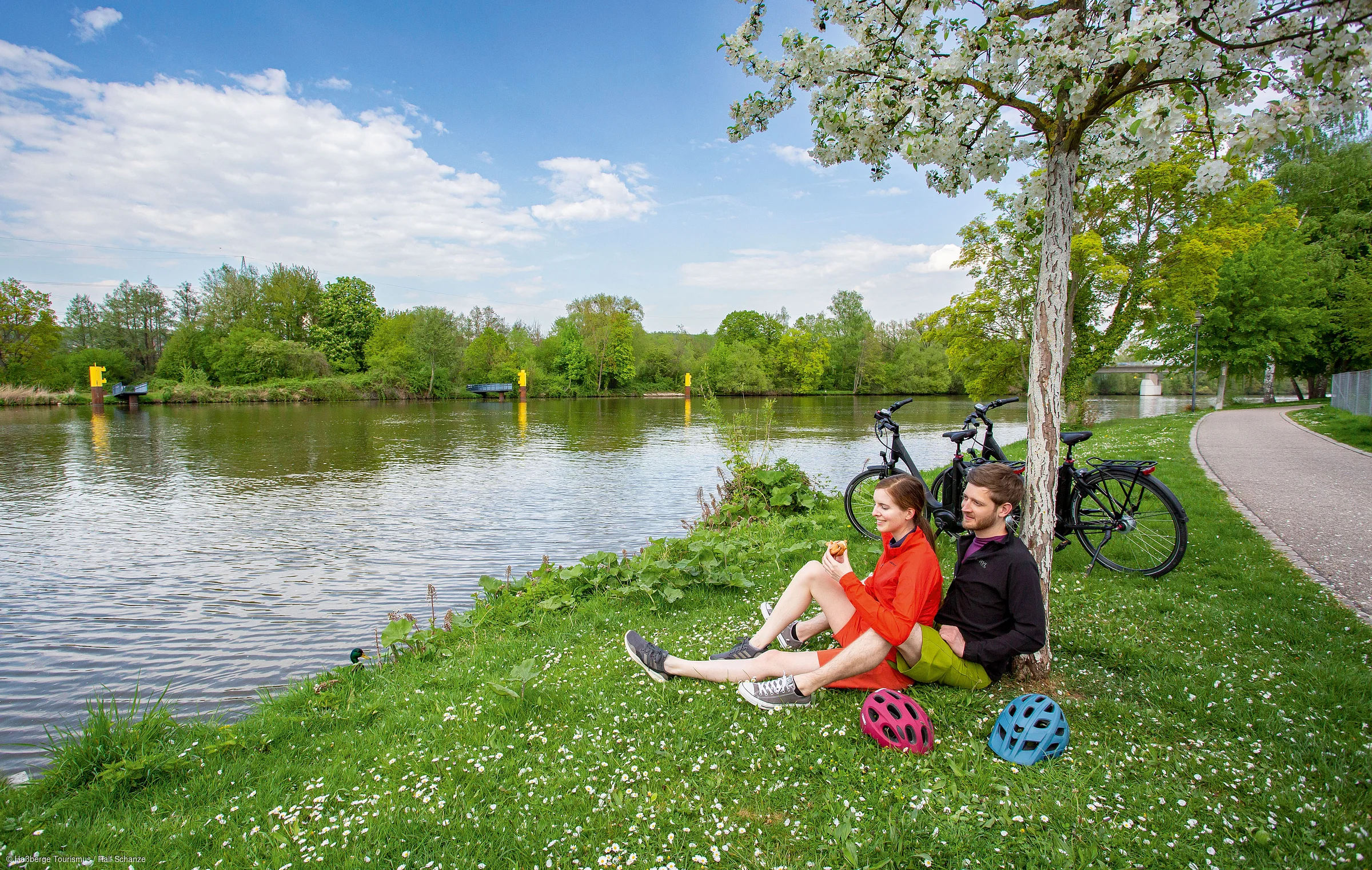Paar sitzt am Flussufer auf Gras unter blühendem Baum, zwei Fahrräder und Helme stehen daneben.