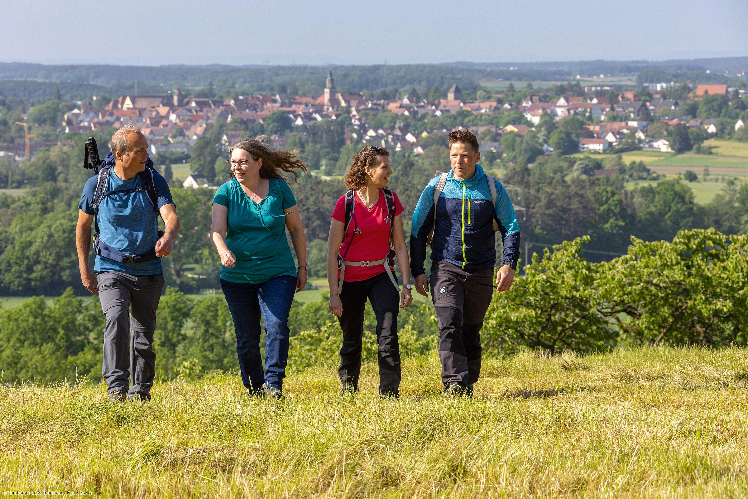 Vier Erwachsene mit Rucksäcken wandern auf einer Wiese vor einer Stadt mit vielen Häusern und Bäumen.