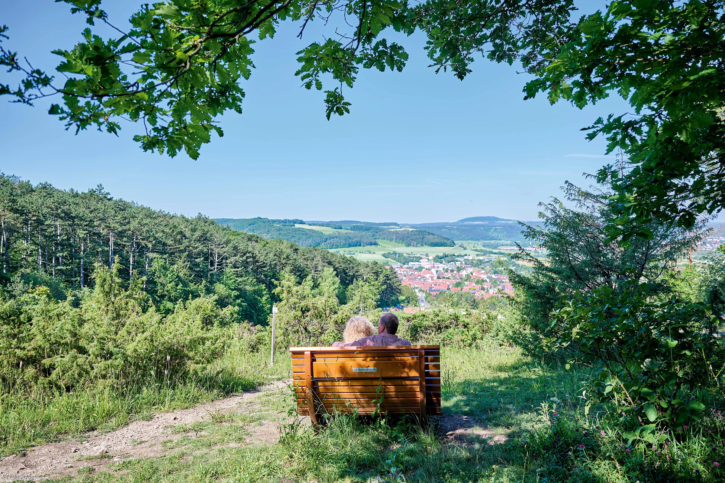 Paar sitzt auf einer Holzbank auf einem Hügel mit Blick auf Wald, Tal und Dorf im Hintergrund.