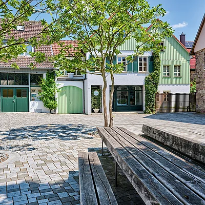 Platz mit gepflastertem Boden, Bänken, Bäumen und Häusern mit grünen Türen und Fensterläden bei Sonnenschein