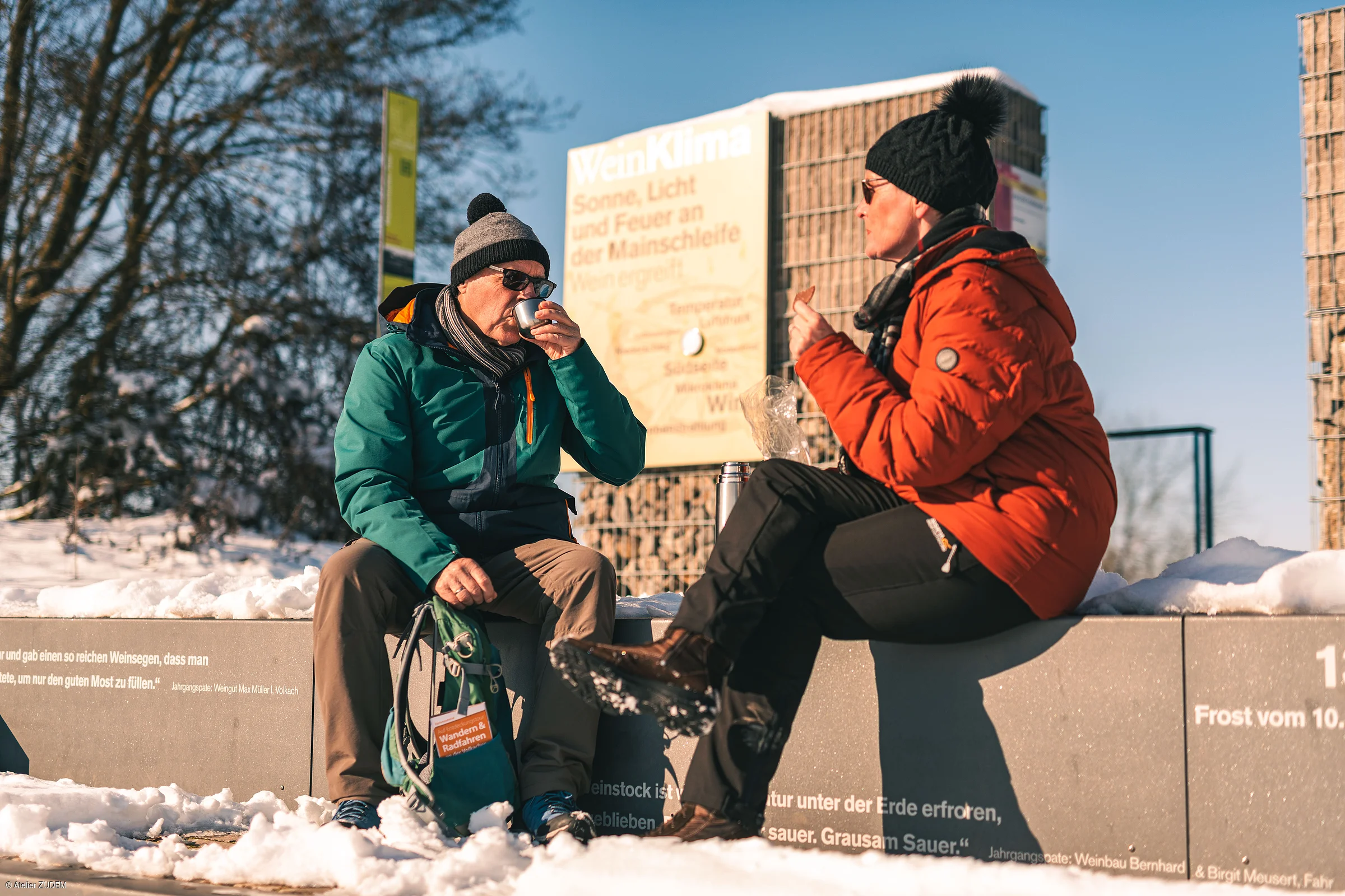 Zwei Personen in Winterkleidung sitzen auf einer Mauer im Schnee und trinken aus Bechern.
