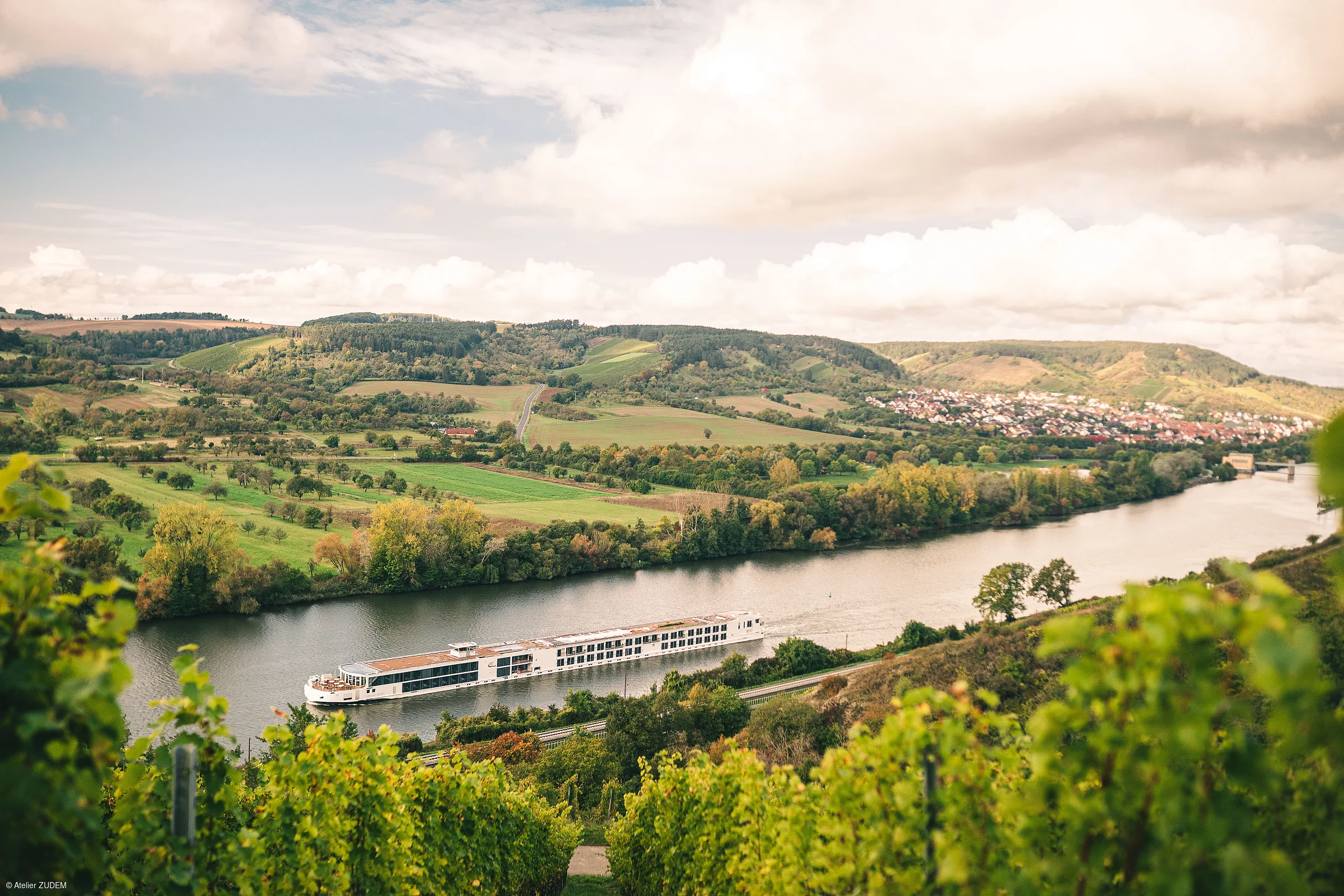 Fluss mit Fahrgastschiff, umgeben von grünen Feldern, Hügeln und einem Dorf im Hintergrund bei bewölktem Himmel.