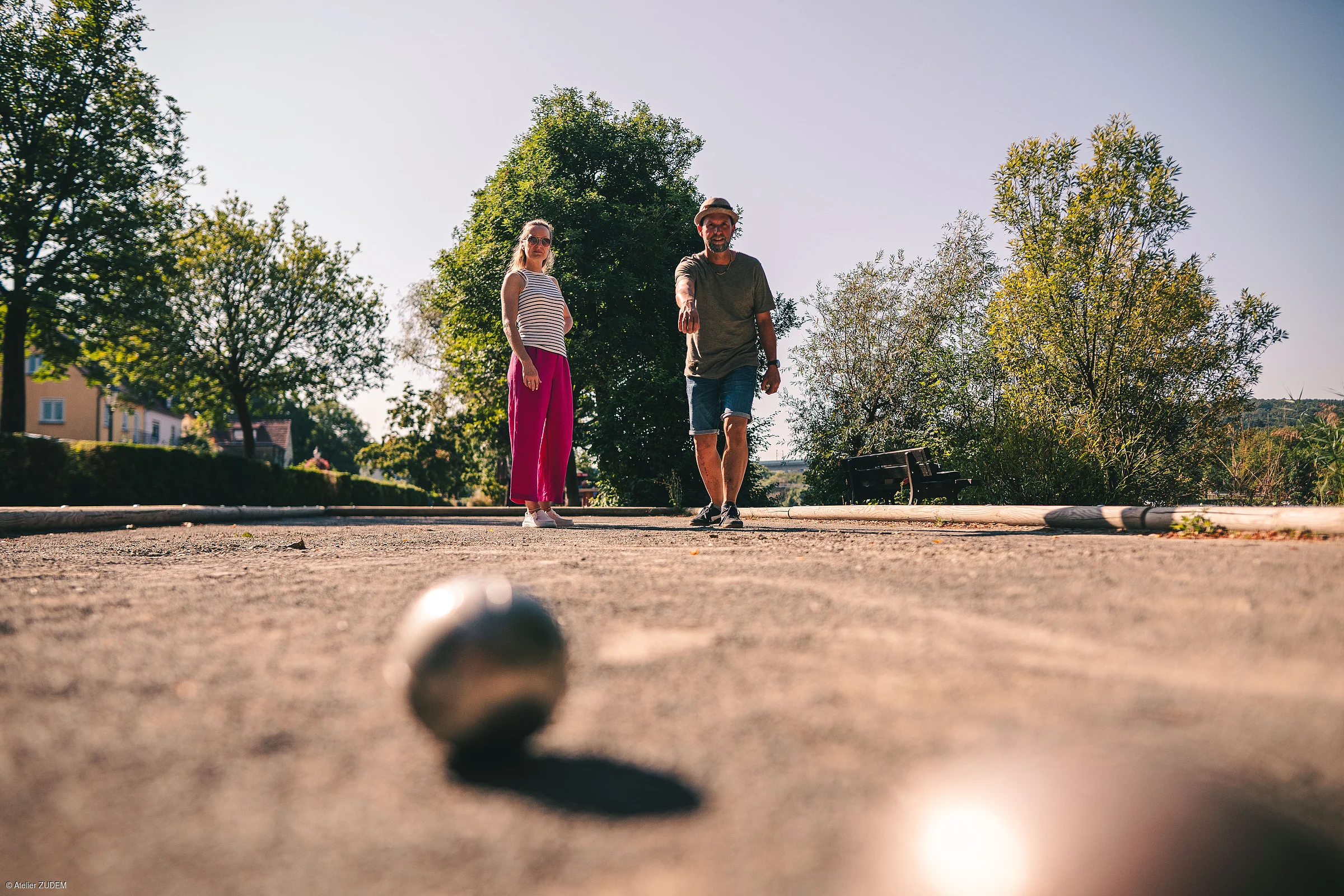 Zwei Personen spielen Boule auf einem Sandplatz im Freien, umgeben von Bäumen und Bänken.