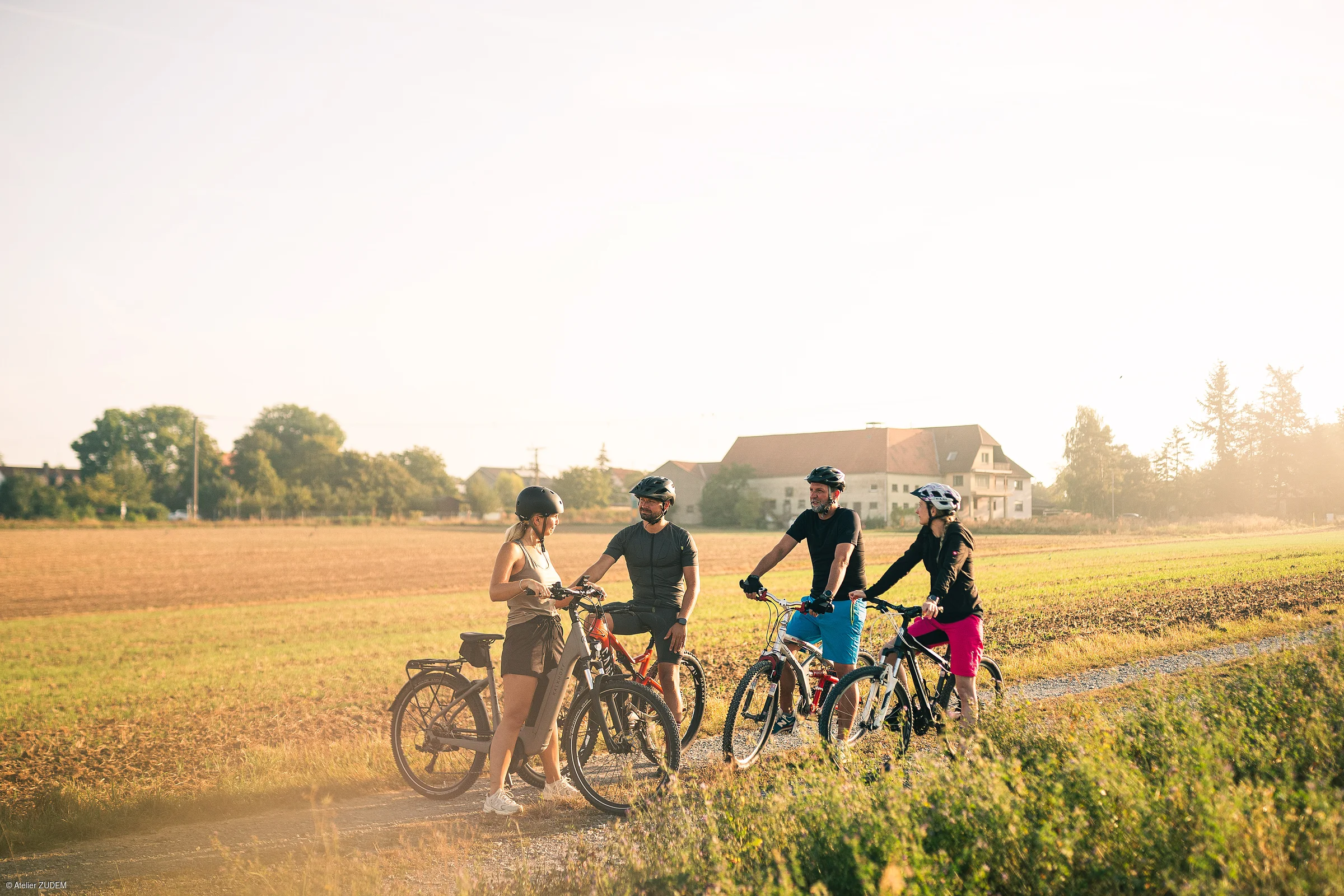 Vier Personen mit Fahrradhelmen stehen mit ihren Fahrrädern auf einem Feldweg in ländlicher Umgebung.