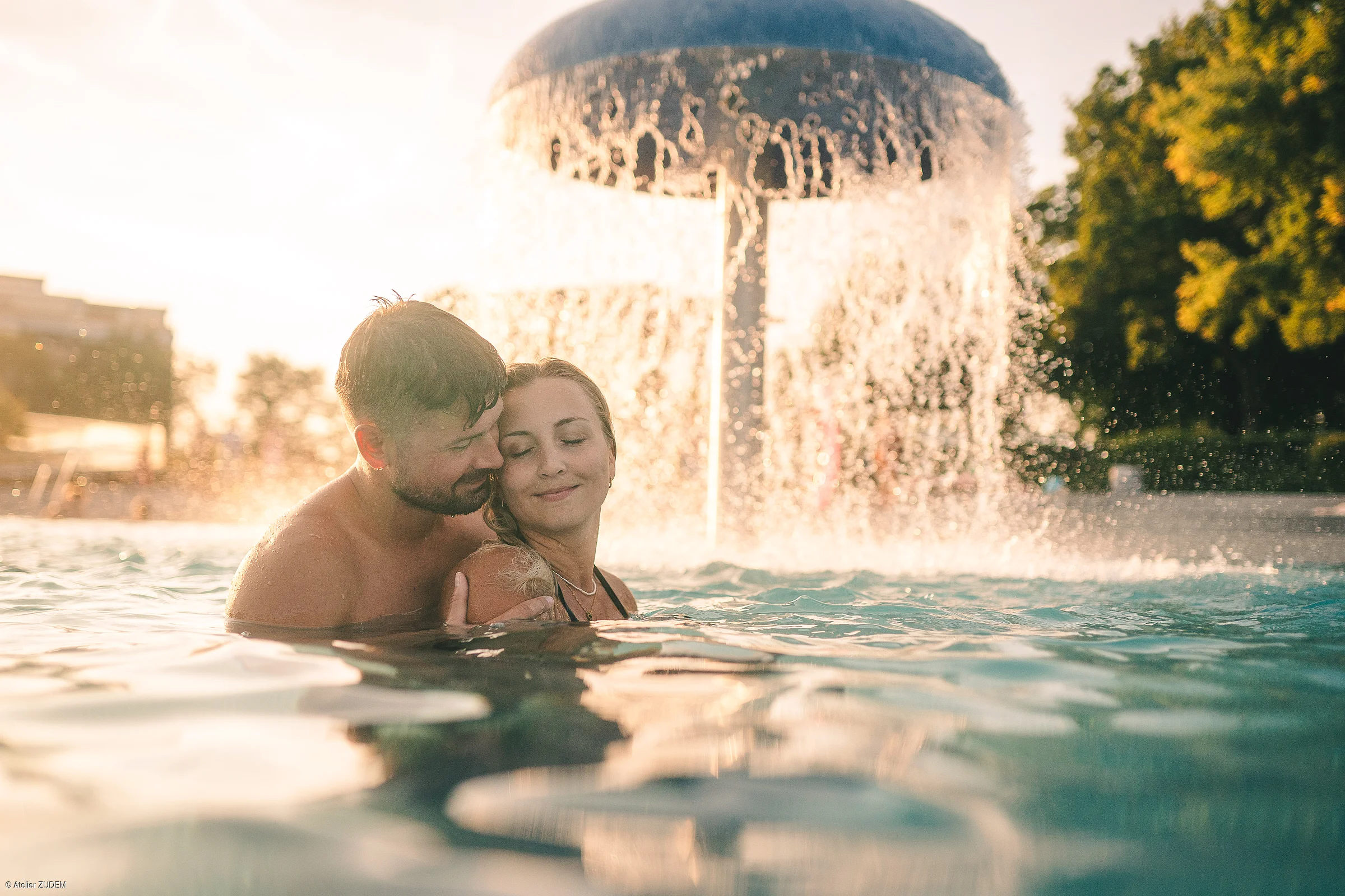 Paar umarmt sich im Wasser vor einem großen Pilzbrunnen bei Sonnenuntergang im Freien.
