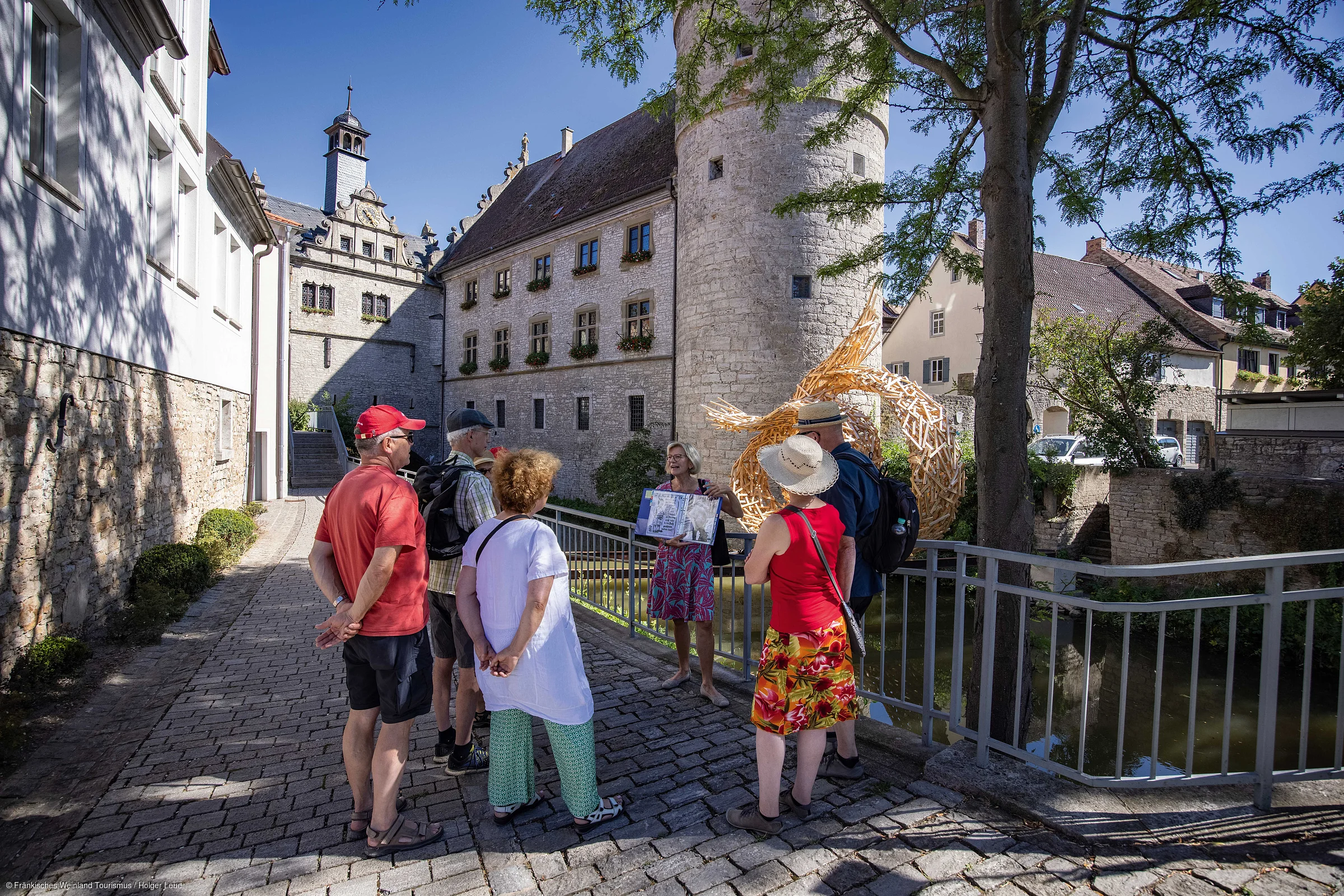 Fünf Personen hören einer Frau zu, die vor einem historischen Gebäude mit Turm eine Führung gibt.