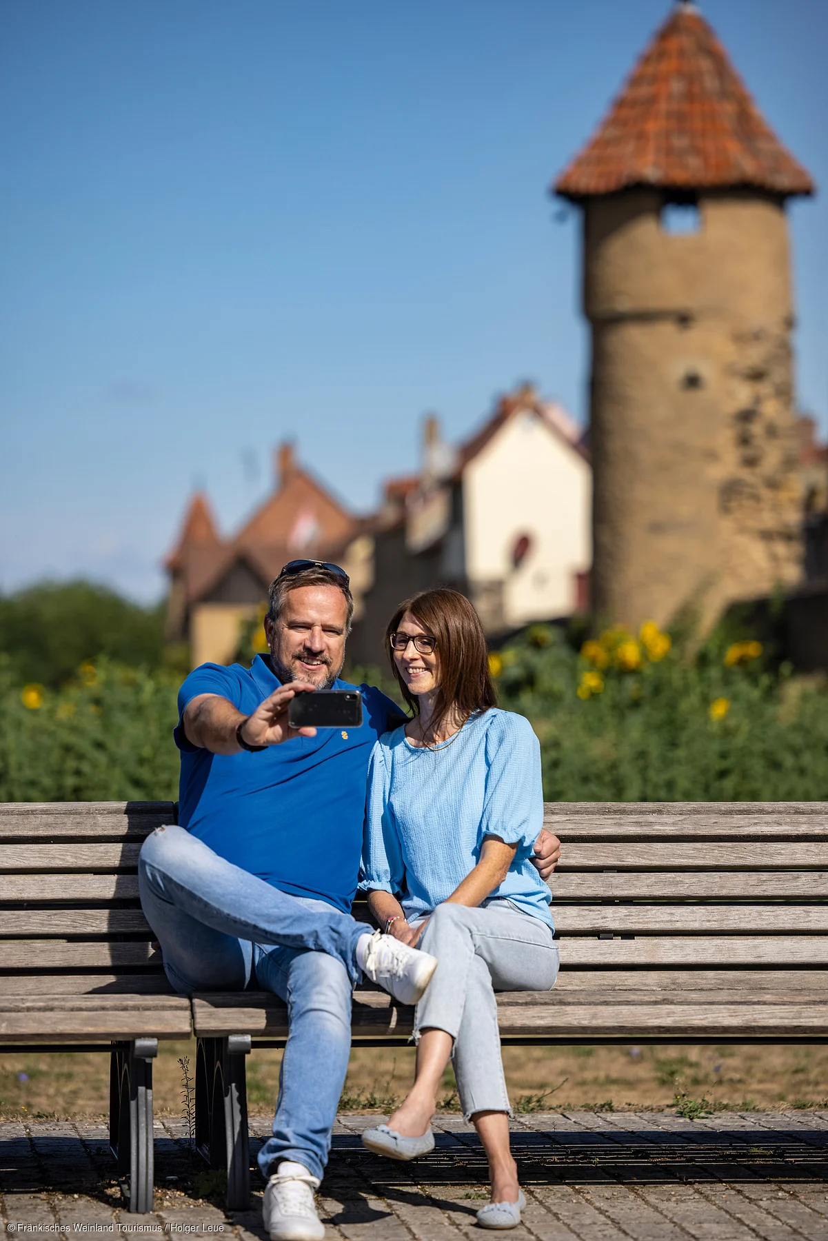 Paar sitzt auf Parkbank vor historischen Gebäuden und macht ein Selfie mit Smartphone.