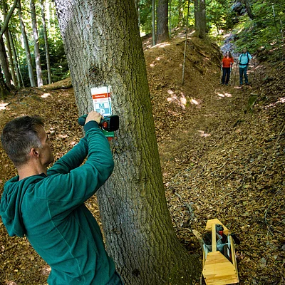 Mann befestigt mit Akkuschrauber ein Schild an einem Baum, zwei Wanderer mit Stöcken auf Waldweg im Hintergrund.