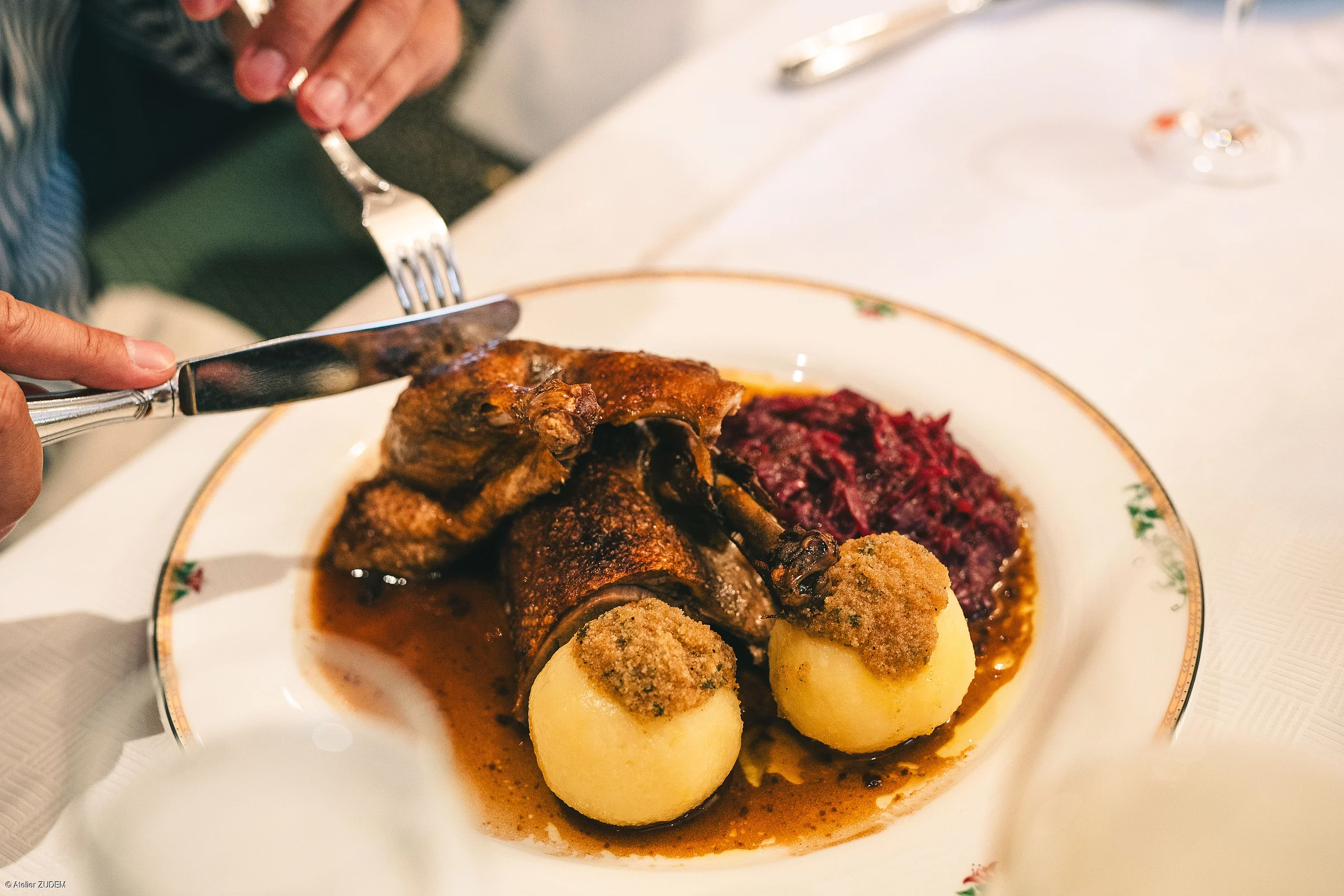 Teller mit gebratener Ente, zwei Klößen mit Bröseln und Rotkohl, Person schneidet mit Messer und Gabel.