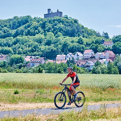 Mann fährt Fahrrad auf Weg vor Feld, im Hintergrund Dorf und bewaldeter Hügel mit Burg.