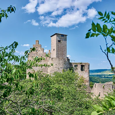 Burgruine mit Turm hinter grünen Bäumen unter blauem Himmel mit Wolken.
