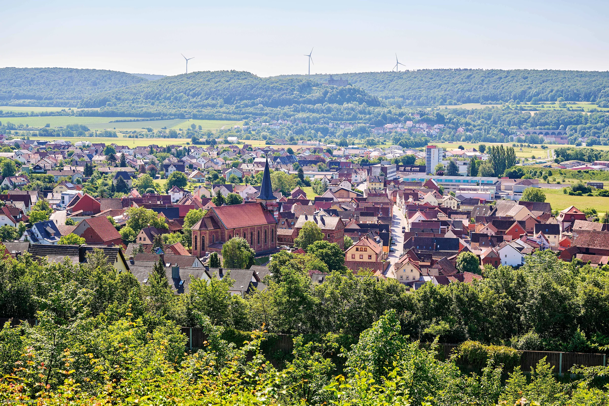 Blick auf eine Kleinstadt mit Kirche, umgeben von Bäumen, Feldern und bewaldeten Hügeln mit Windrädern im Hintergrund.