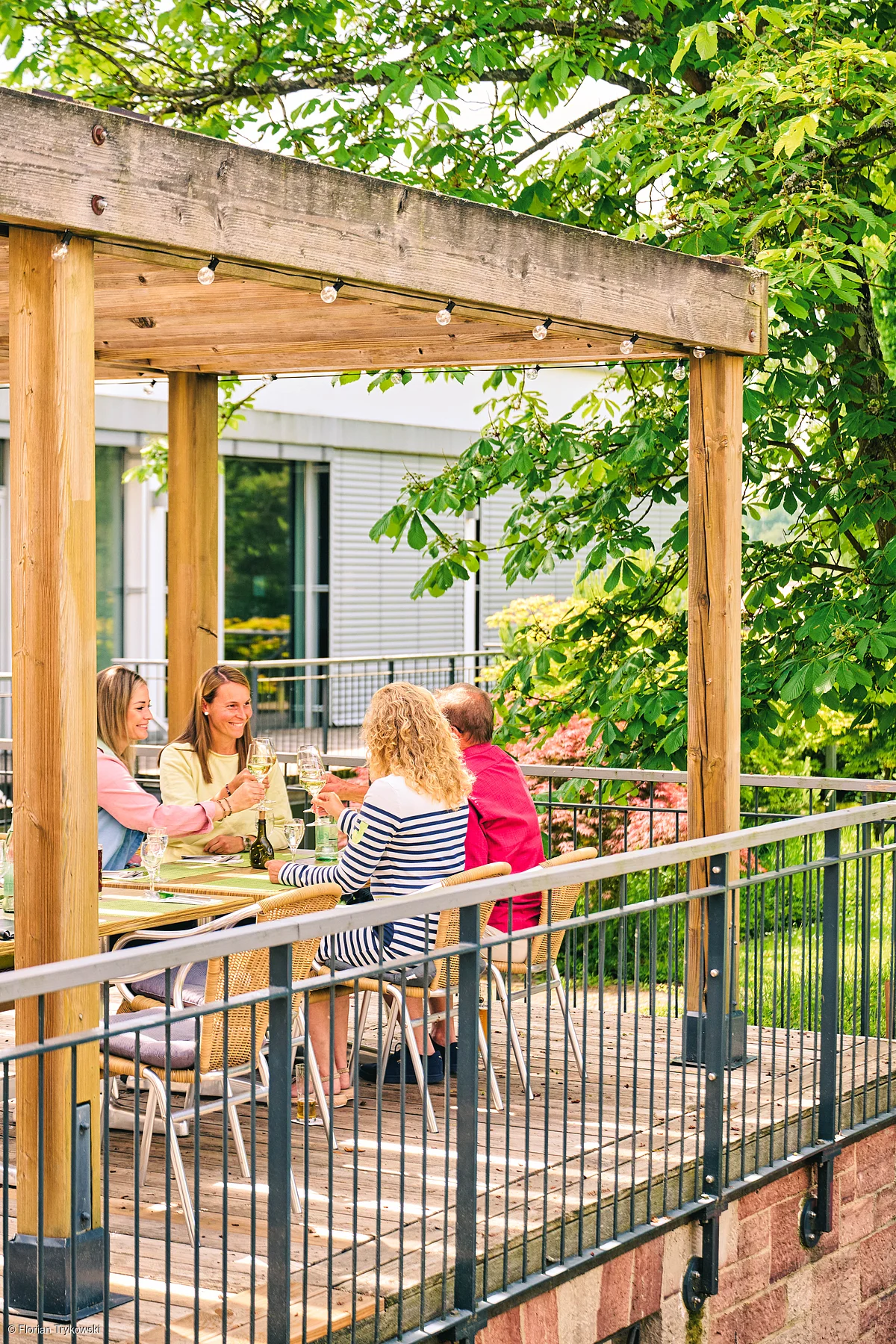 Vier Personen sitzen an einem Holztisch unter einem Holzpavillon und stoßen mit Weingläsern an.