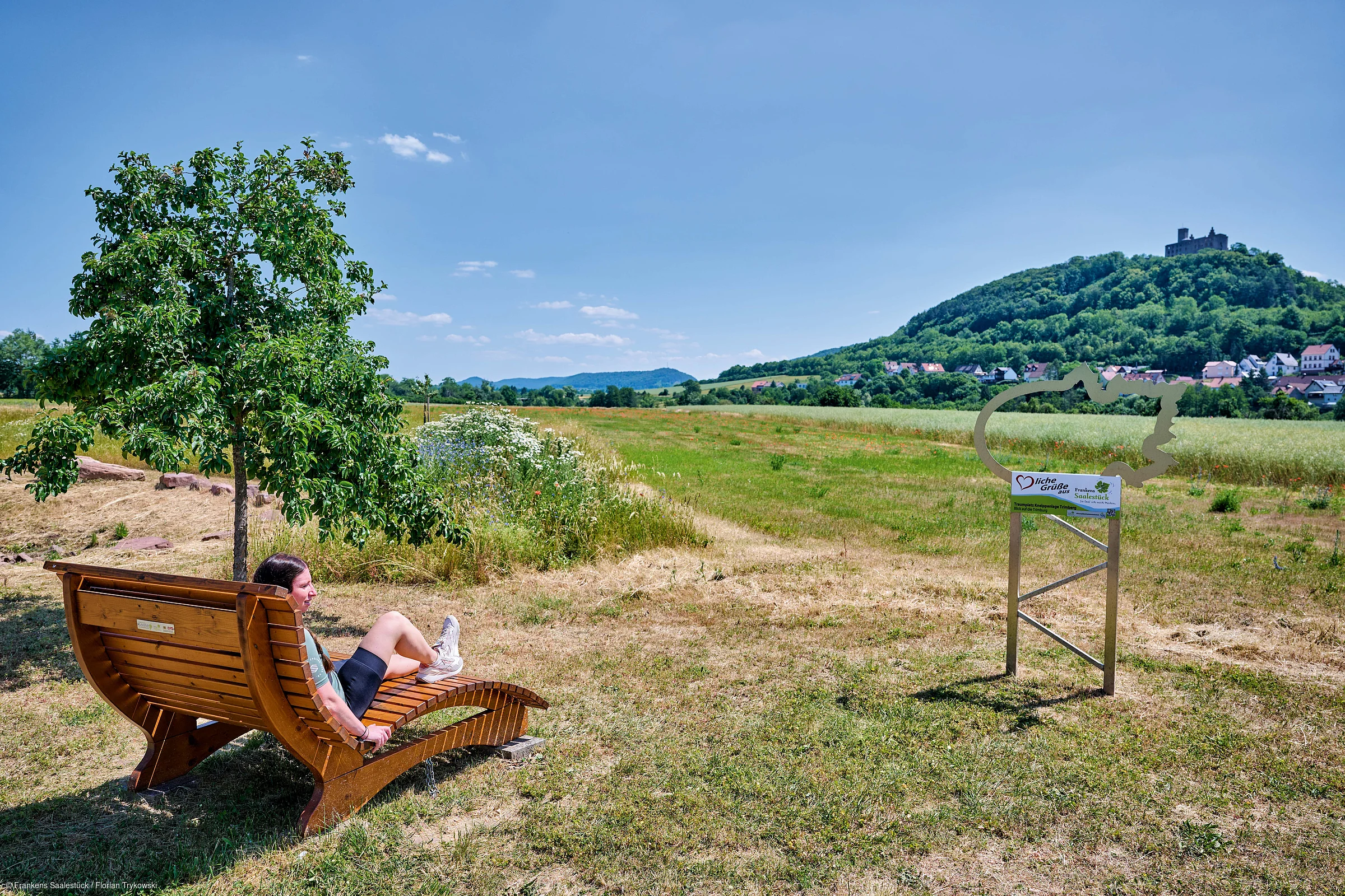 Frau sitzt auf Holzliege unter Baum auf Wiese, im Hintergrund Hügel mit Burg und Ort bei klarem Himmel.