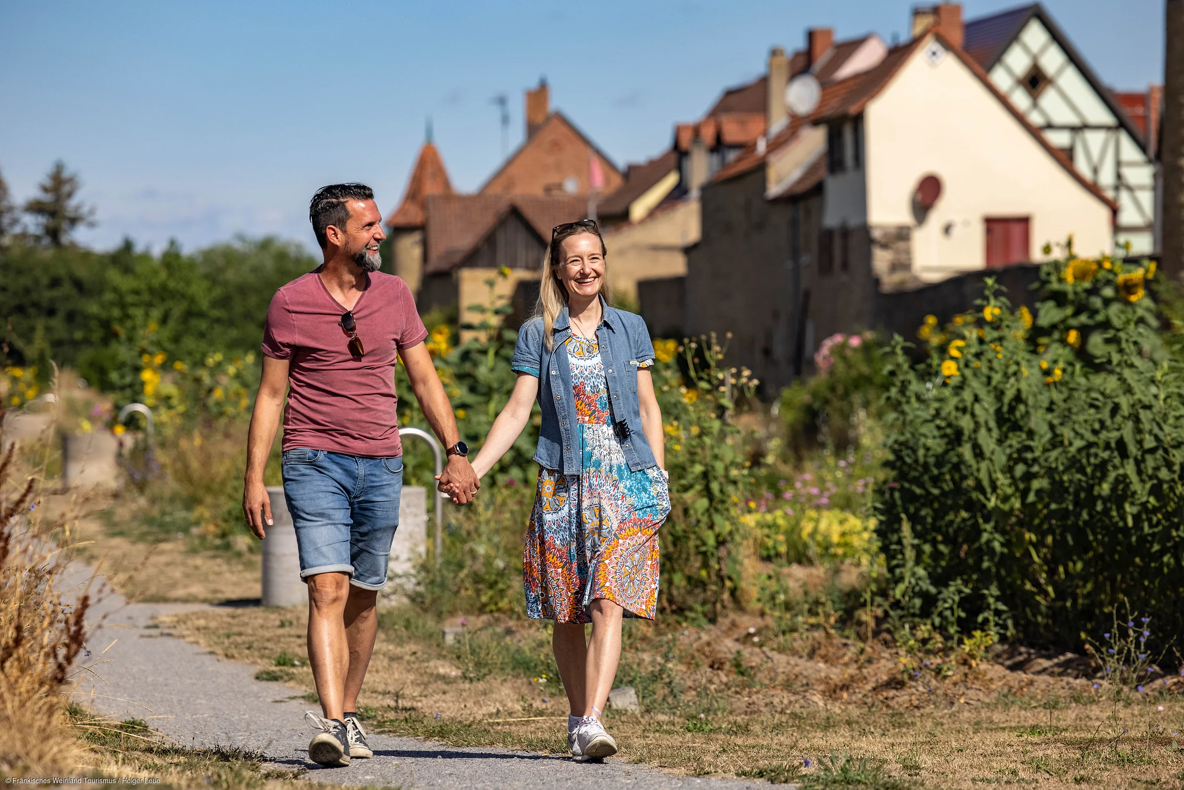 Paar hält Händchen und spaziert auf einem Weg mit Blumen und Häusern im Hintergrund bei Sonnenschein.