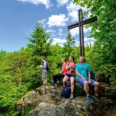 Vier Wanderer sitzen und stehen auf Felsen neben einem großen Holzkreuz in einem bewaldeten Gebiet.