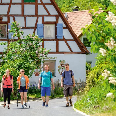 Vier Personen wandern auf einer Straße vor einem Fachwerkhaus mit blühenden Büschen im Sommer.