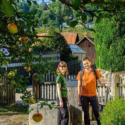 Zwei Wanderer mit Trekkingstöcken stehen auf einem Kiesweg vor einem Holzzaun und grünen Büschen im Dorf.