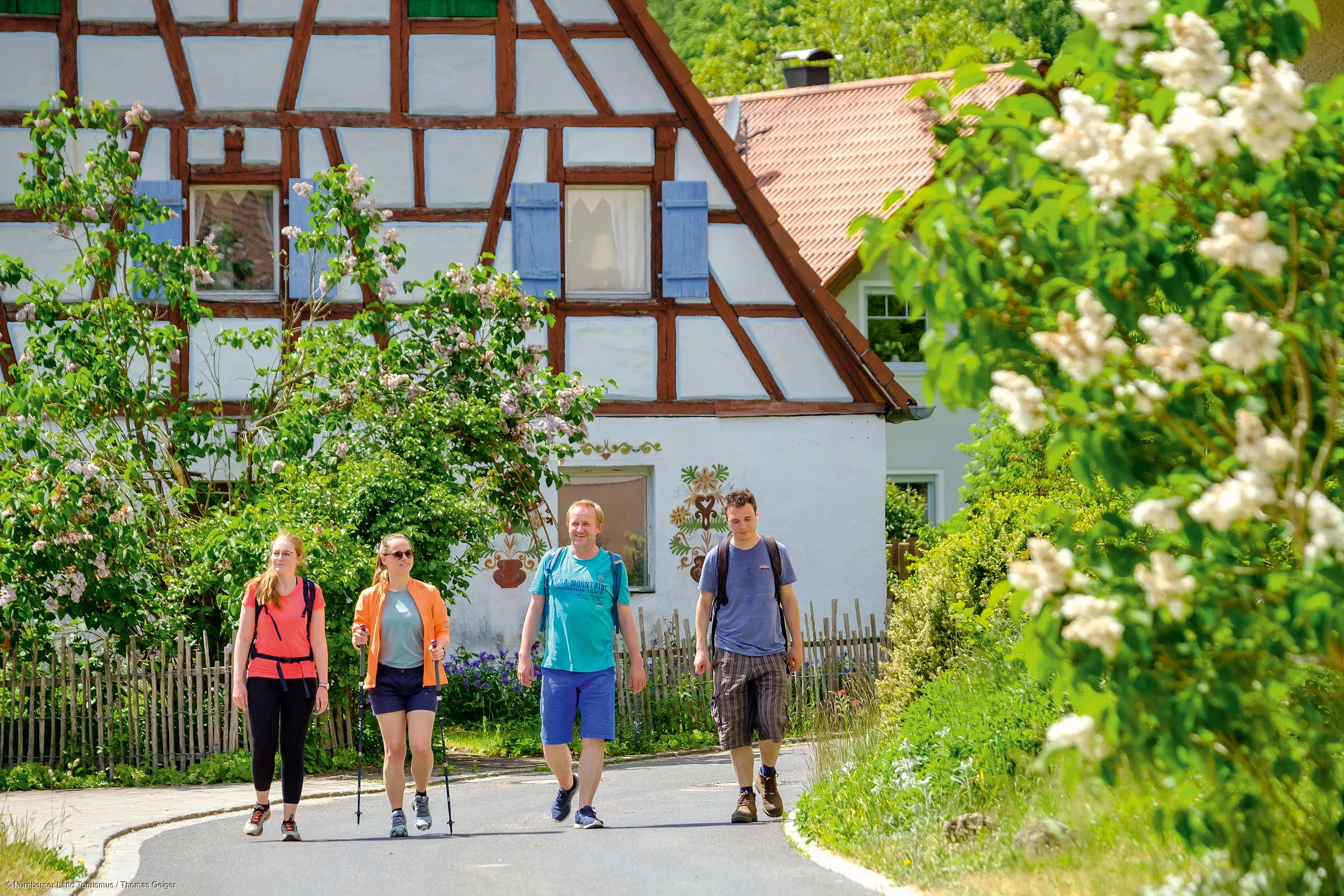 Vier Personen wandern auf einer Straße vor einem Fachwerkhaus mit blühenden Büschen im Sommer.