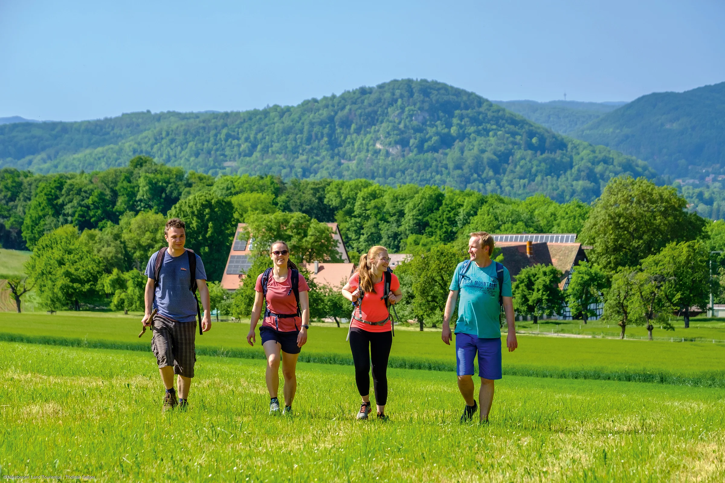 Vier Personen mit Rucksäcken wandern auf einer grünen Wiese vor bewaldeten Hügeln und Häusern.