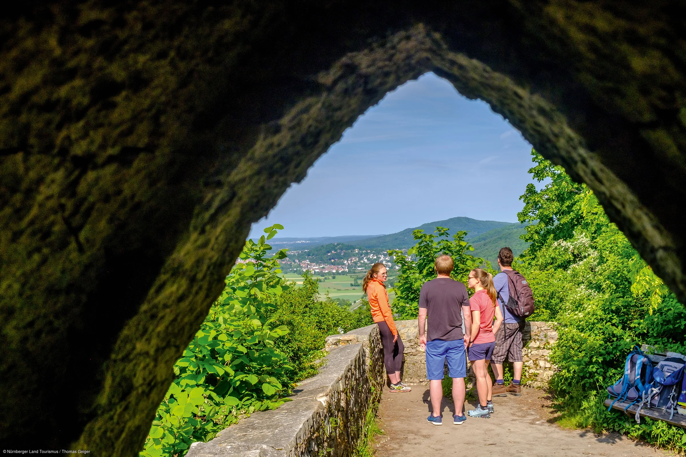 Vier Personen stehen auf einem Weg mit Steinmauer, umgeben von grünen Bäumen, Blick auf Landschaft und Berge.