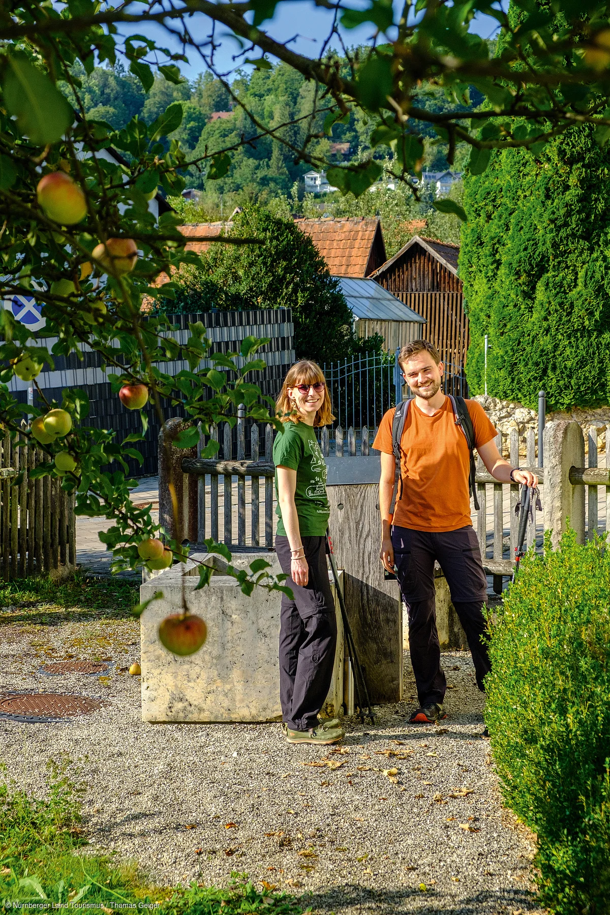 Zwei Wanderer mit Trekkingstöcken stehen auf einem Kiesweg vor einem Holzzaun und grünen Büschen im Dorf.