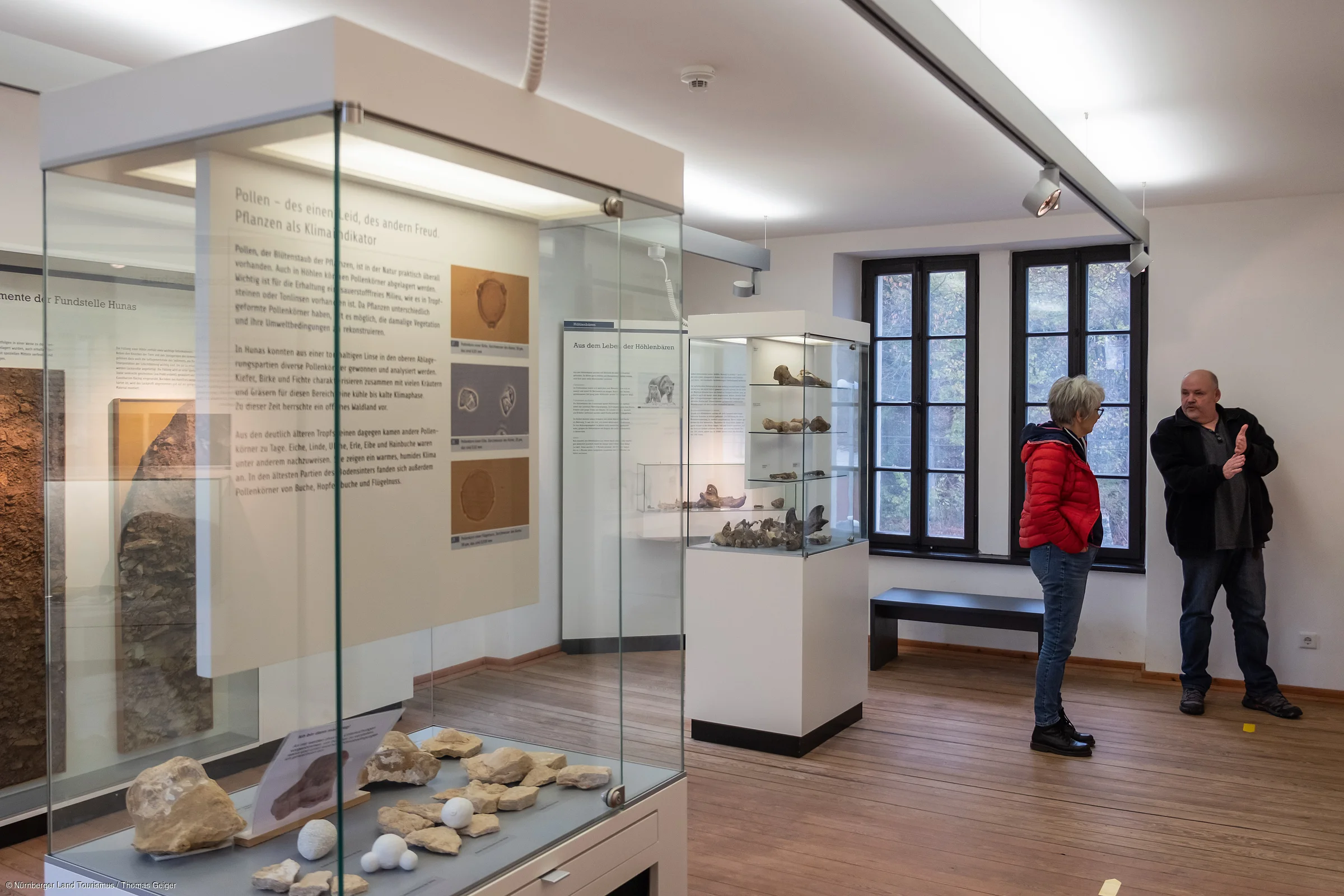 Museumsausstellung mit Vitrinen, Exponaten und zwei Personen im Gespräch vor Fenstern in einem Raum mit Holzboden.