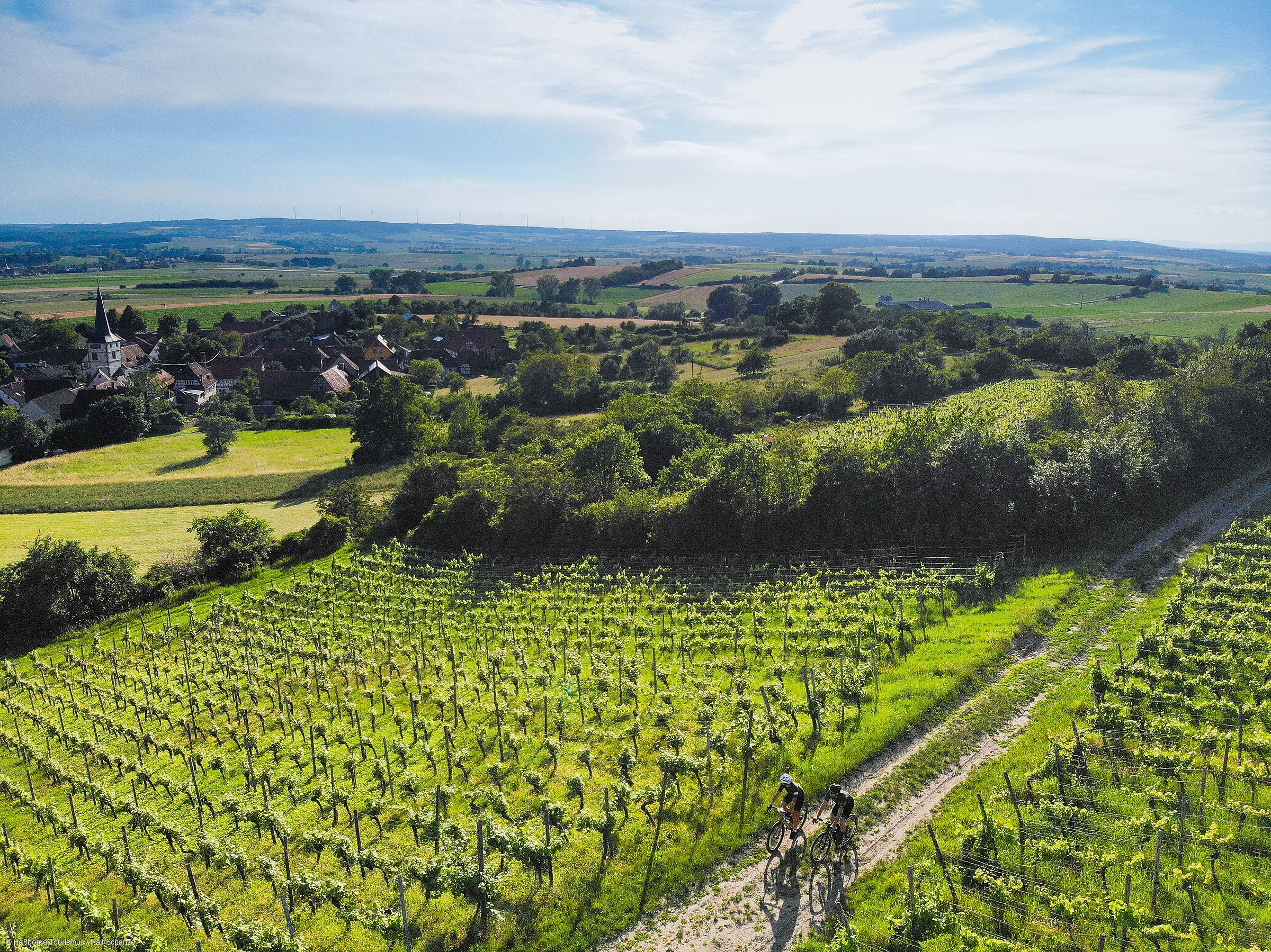 Zwei Radfahrer auf Weg durch grüne Weinberge mit Dorf und Hügeln im Hintergrund unter blauem Himmel.
