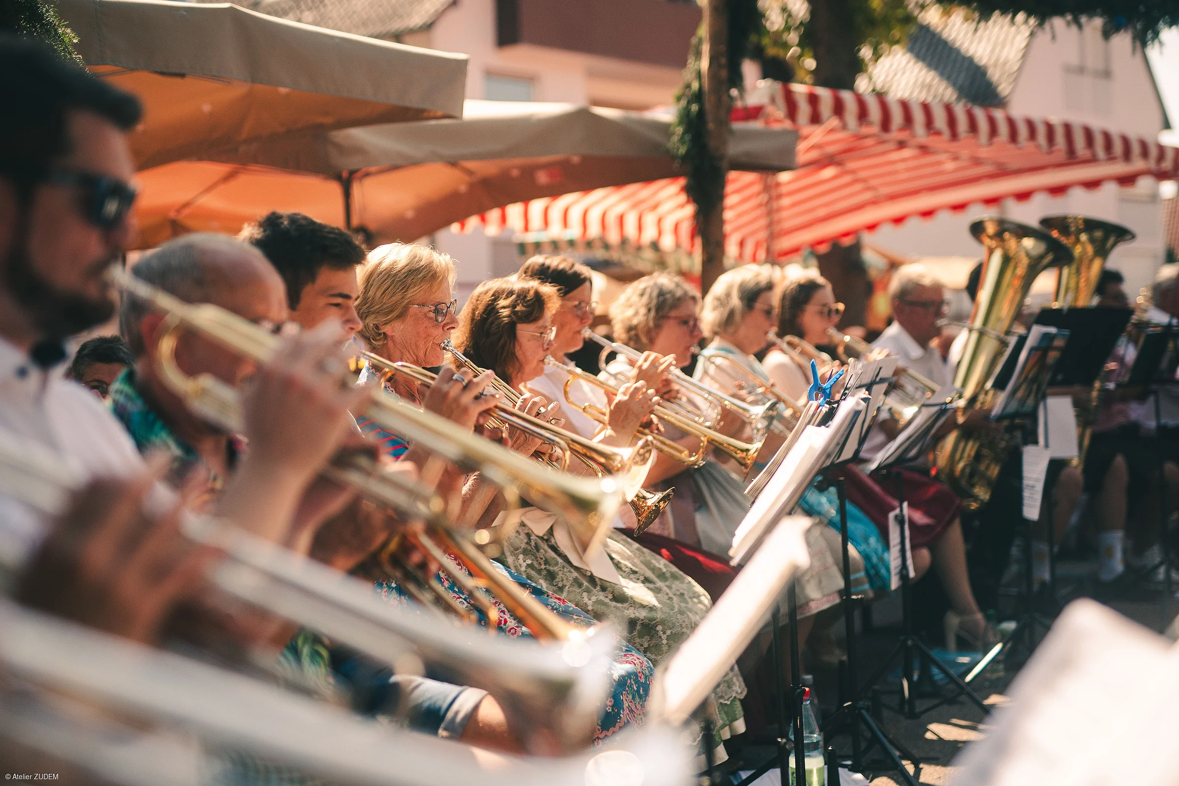 Blasmusikgruppe spielt Trompeten und Tubas bei sonnigem Freiluftkonzert unter Markisen und Sonnenschirmen.