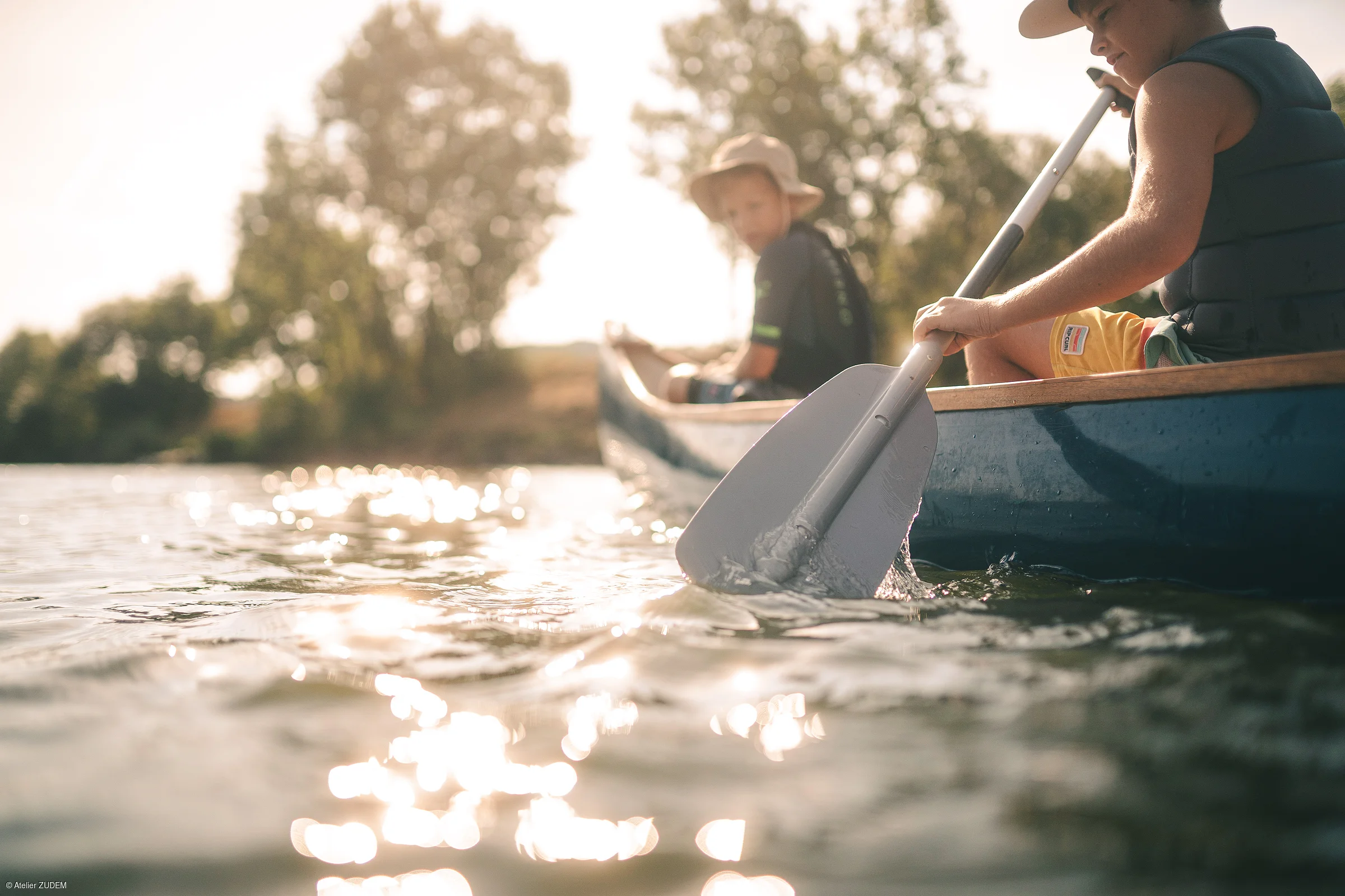 Zwei Kinder mit Hüten paddeln im Kanu auf einem sonnigen Gewässer, Bäume im Hintergrund.