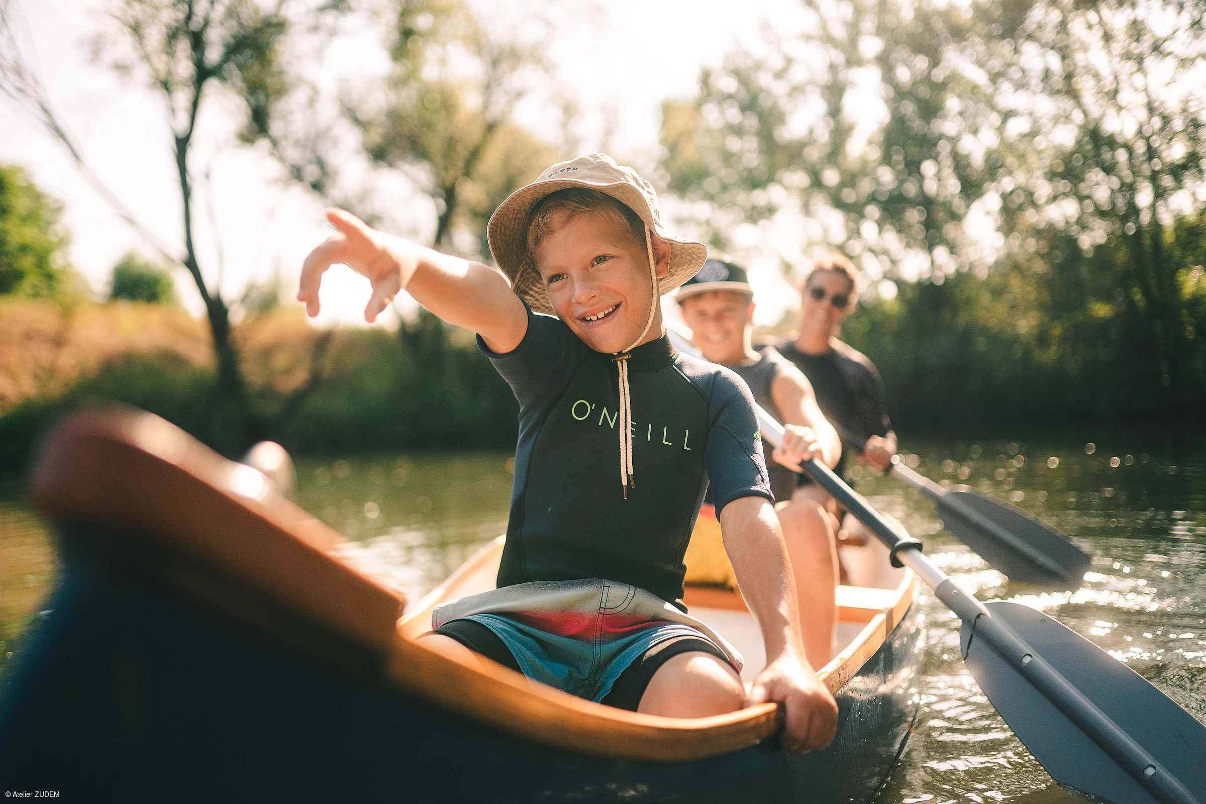 Drei Personen in einem Kanu auf einem Fluss, vorne ein Junge mit Sonnenhut, der nach vorne zeigt.