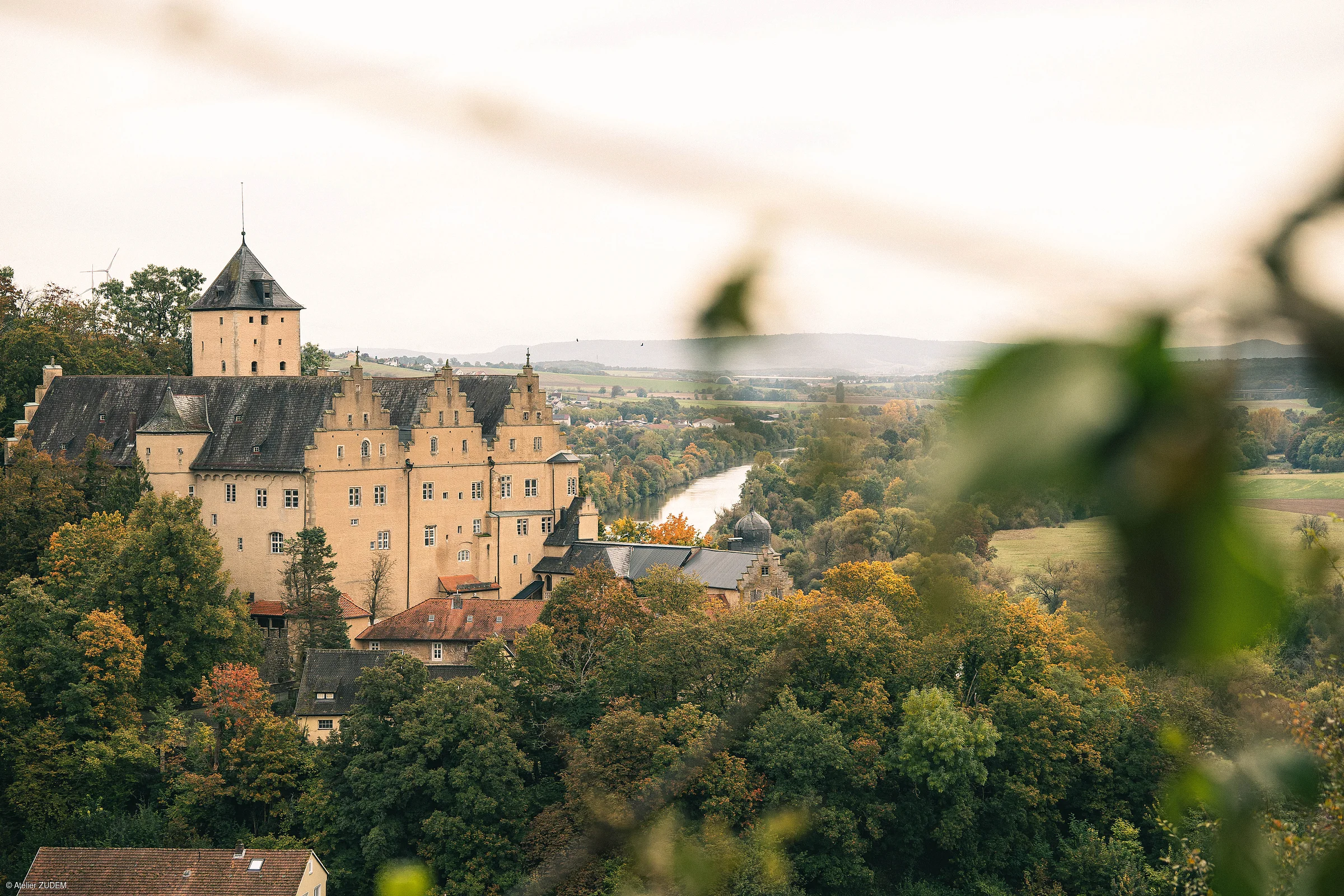 Blick auf ein Schloss mit Turm, umgeben von Bäumen und einem Fluss im Hintergrund, im Herbst.
