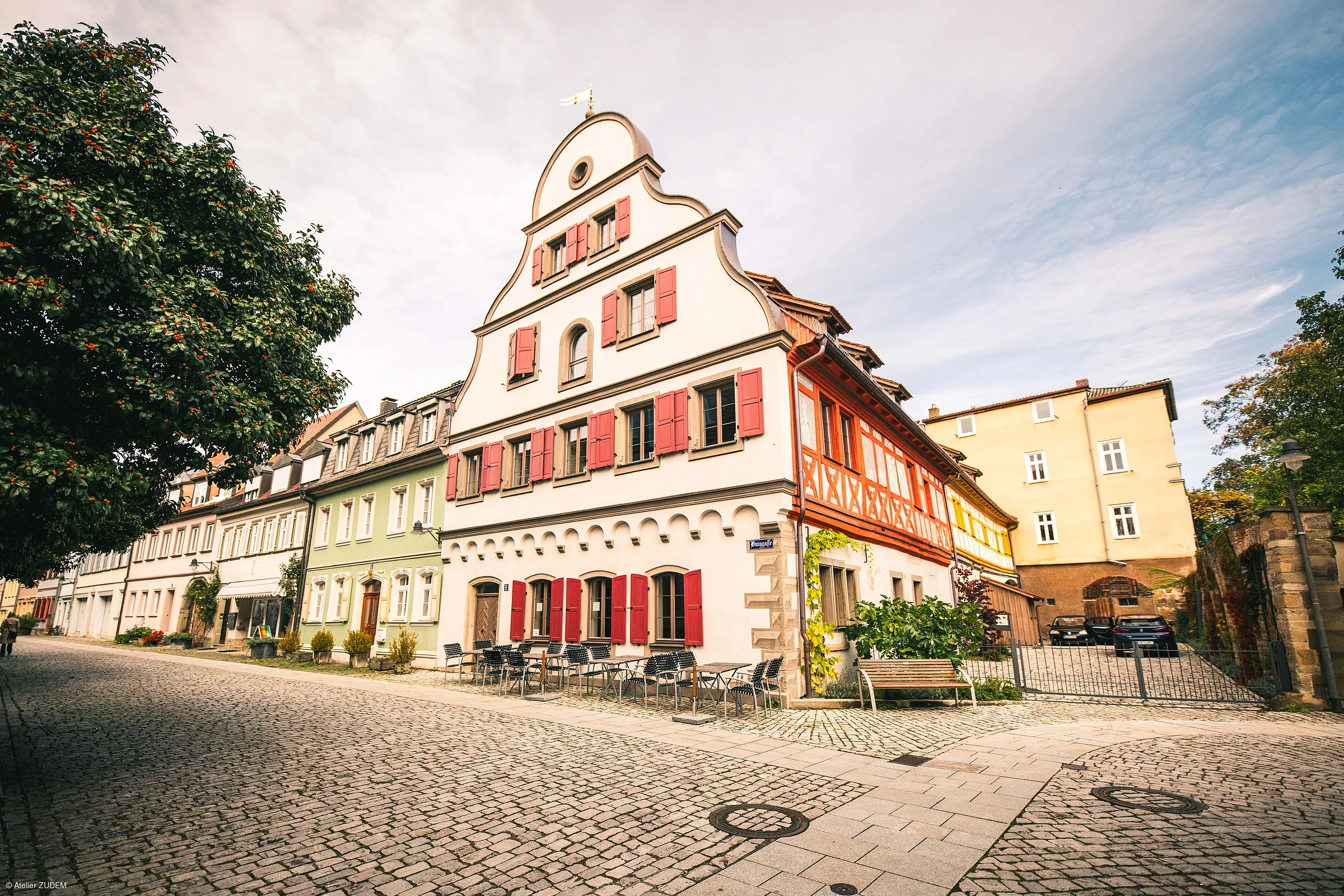 Historische Fachwerkhäuser mit roten Fensterläden an gepflasterter Straße unter bewölktem Himmel.