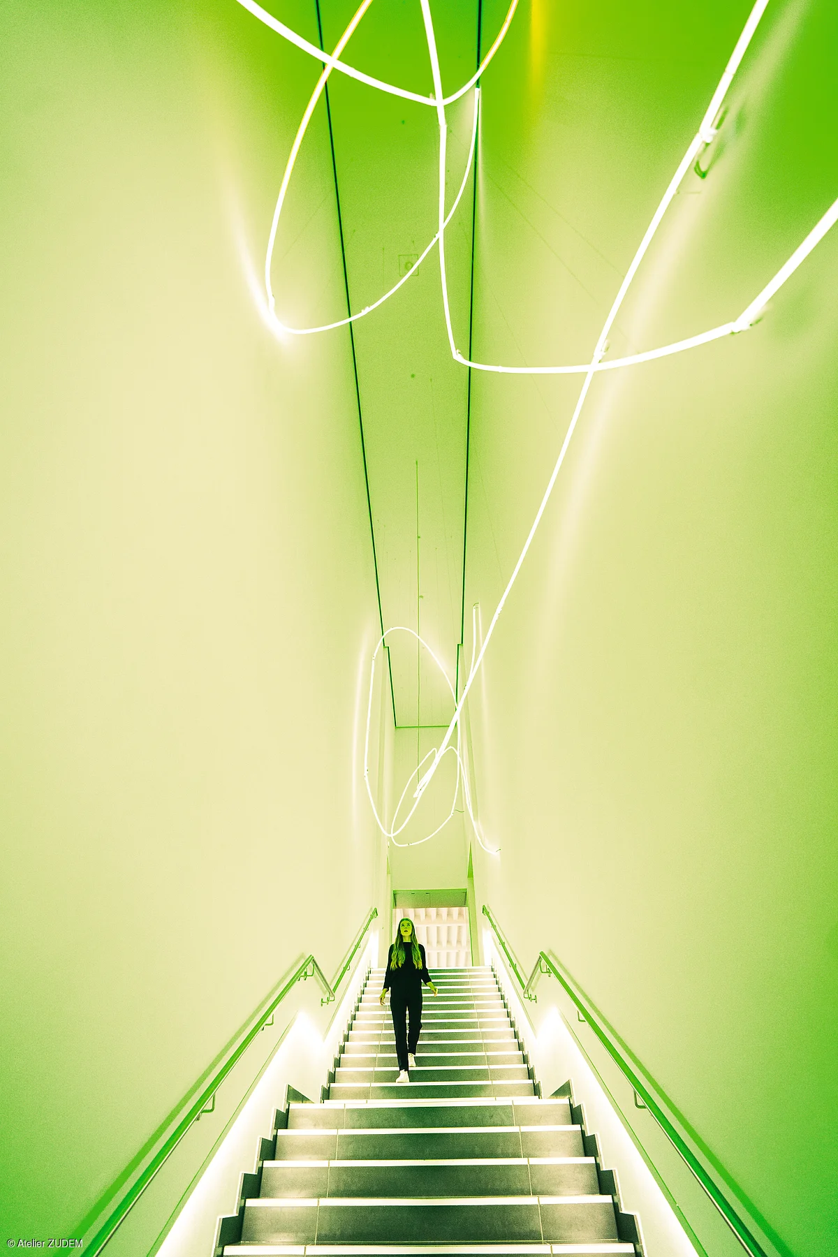 Frau geht eine beleuchtete Treppe in einem grünen, schmalen Raum mit weißen Neonlichtern an der Decke hinunter.