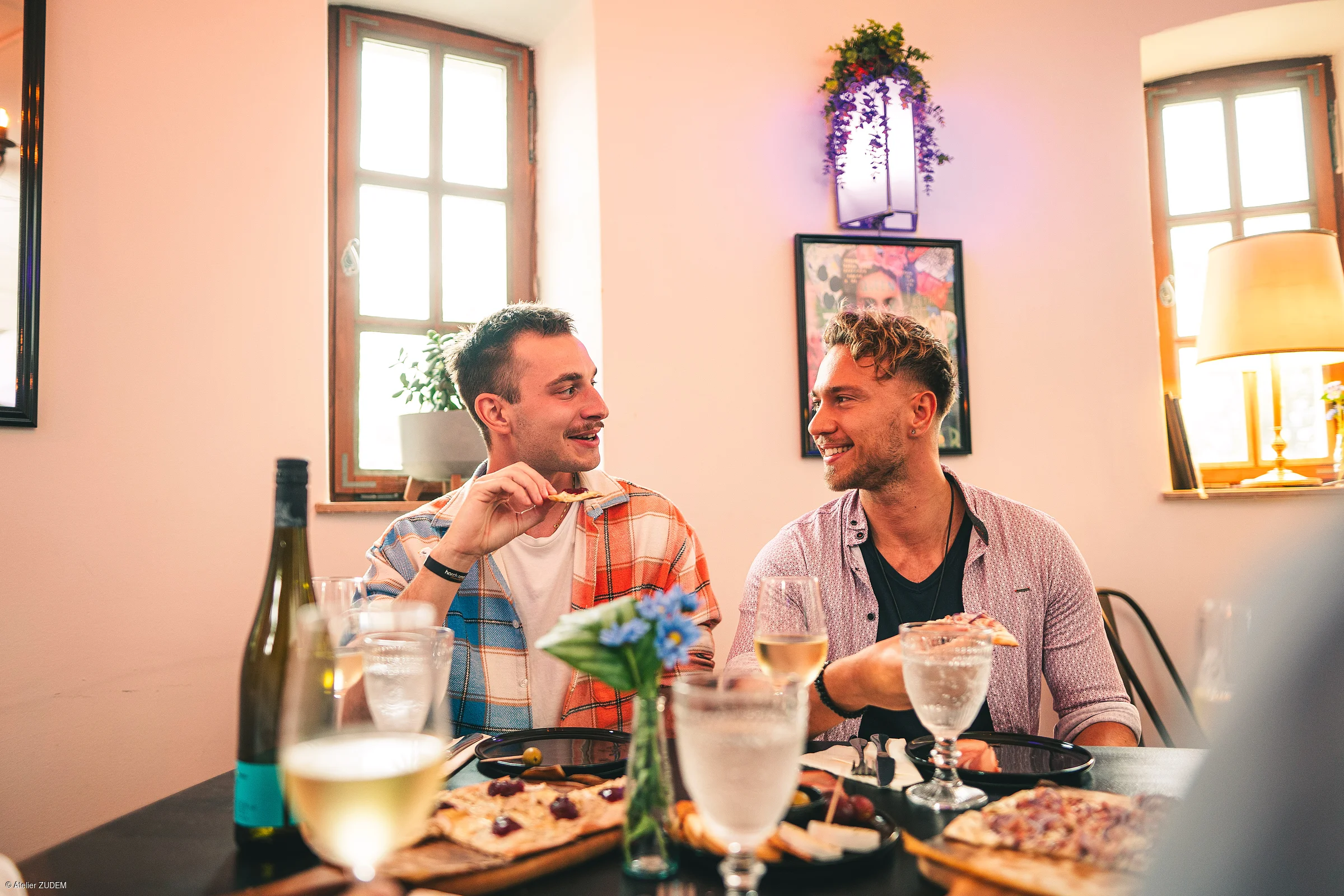 Zwei Männer sitzen an einem Tisch mit Essen und Getränken und unterhalten sich in einem hellen Raum.