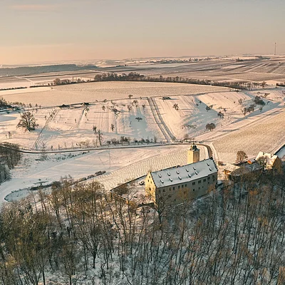 Luftaufnahme eines schneebedeckten Gebäudes in einem winterlichen Waldgebiet mit Feldern im Hintergrund.