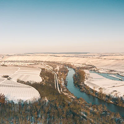 Landschaft mit Fluss, schneebedeckten Feldern und Wald unter klarem Himmel im Winter.