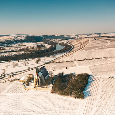 Luftaufnahme einer Kirche umgeben von schneebedeckten Feldern und einem Fluss im Hintergrund bei klarem Himmel.
