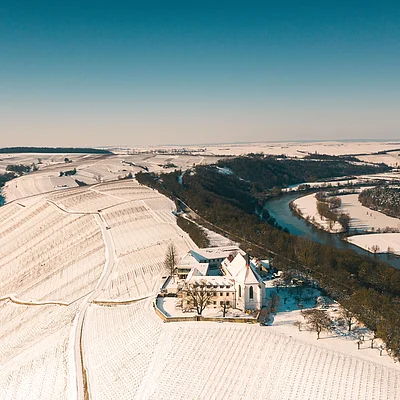Luftaufnahme einer verschneiten Landschaft mit Fluss, Feldern und Gebäuden unter klarem Himmel.