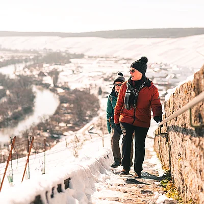 Zwei Personen in Winterkleidung gehen einen schneebedeckten Weg neben einer Steinmauer bergauf.