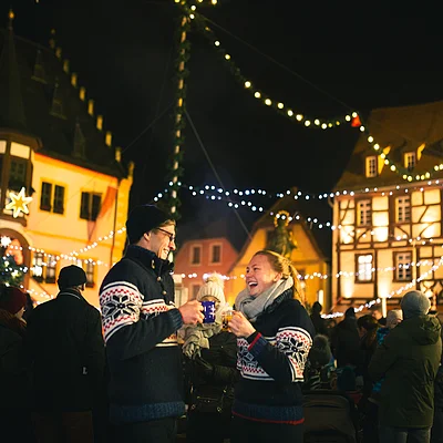 Paar in Winterkleidung trinkt Glühwein auf einem beleuchteten Weihnachtsmarkt bei Nacht.