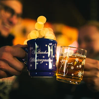 Zwei Personen stoßen mit Tassen an, eine blaue und eine transparente mit Wintermotiven, unscharfer Hintergrund.