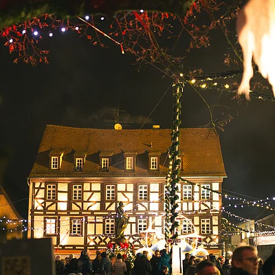 Beleuchtetes Fachwerkhaus bei Nacht mit Menschen und Weihnachtsbeleuchtung auf einem Markt.