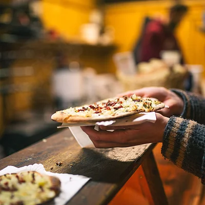 Person hält eine belegte Flammkuchen-Scheibe auf Serviette über einem Holztisch in einer warm beleuchteten Umgebung