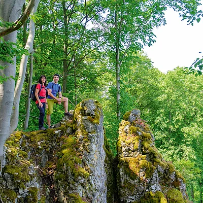 Zwei Wanderer mit Rucksack stehen auf moosbedecktem Felsen im Wald bei Tageslicht.