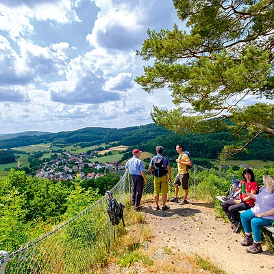 Wandergruppe auf Aussichtsplattform mit Blick auf Tal, Dorf und bewaldete Hügel unter bewölktem Himmel.